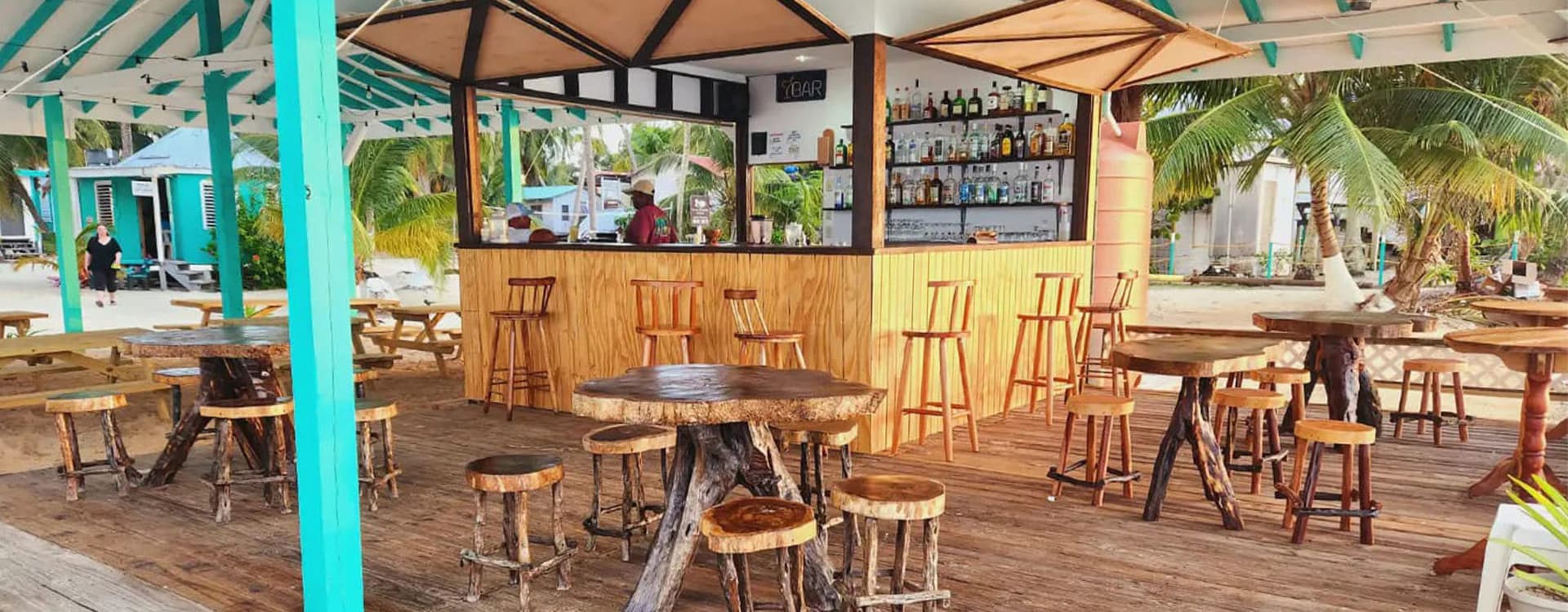 A wooden bar with high stools under a tropical canopy, surrounded by palm trees and rustic tables.
