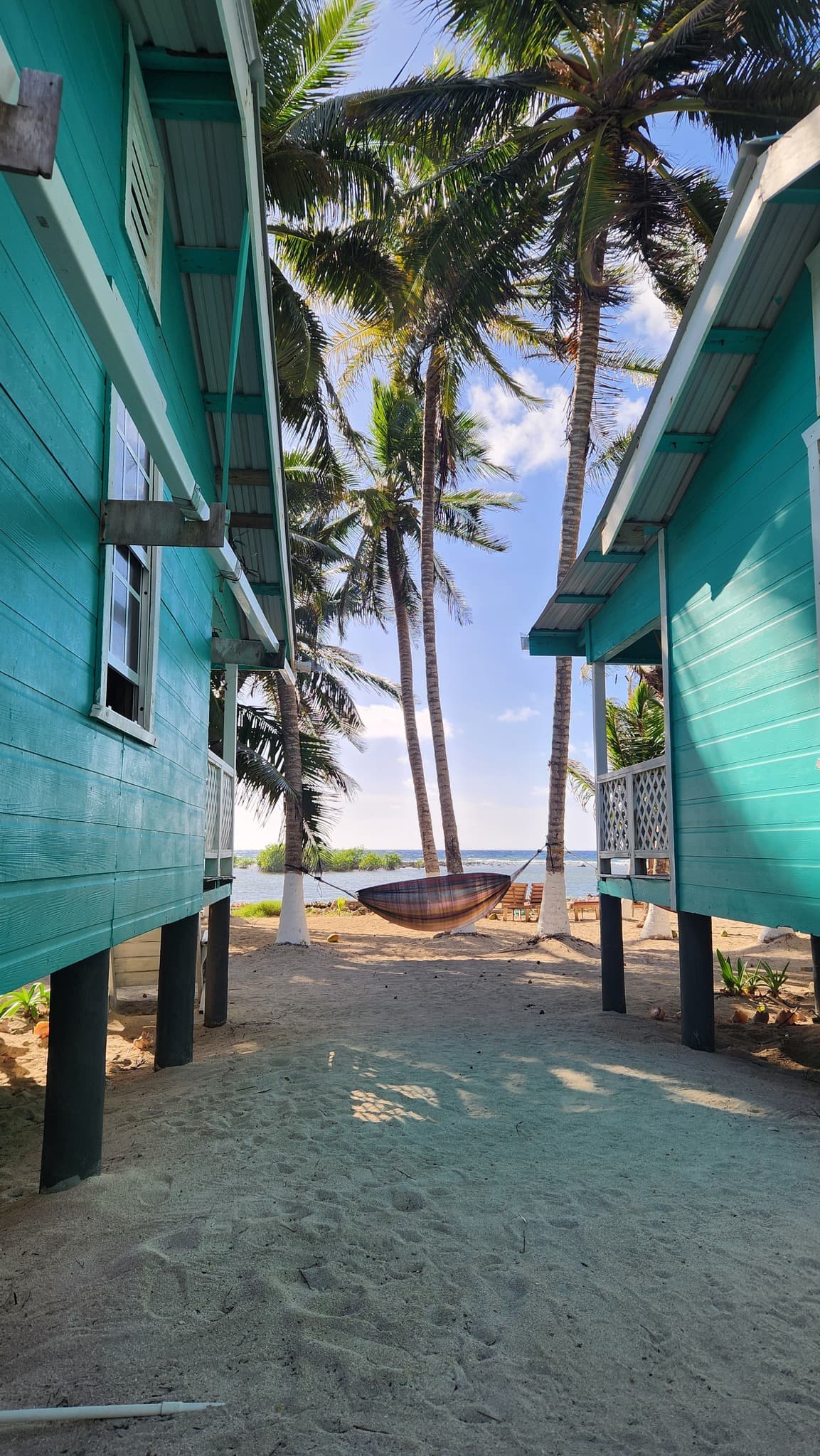 A tranquil beach scene framed by turquoise cottages and palm trees, featuring a hammock and sandy path leading to the ocean.