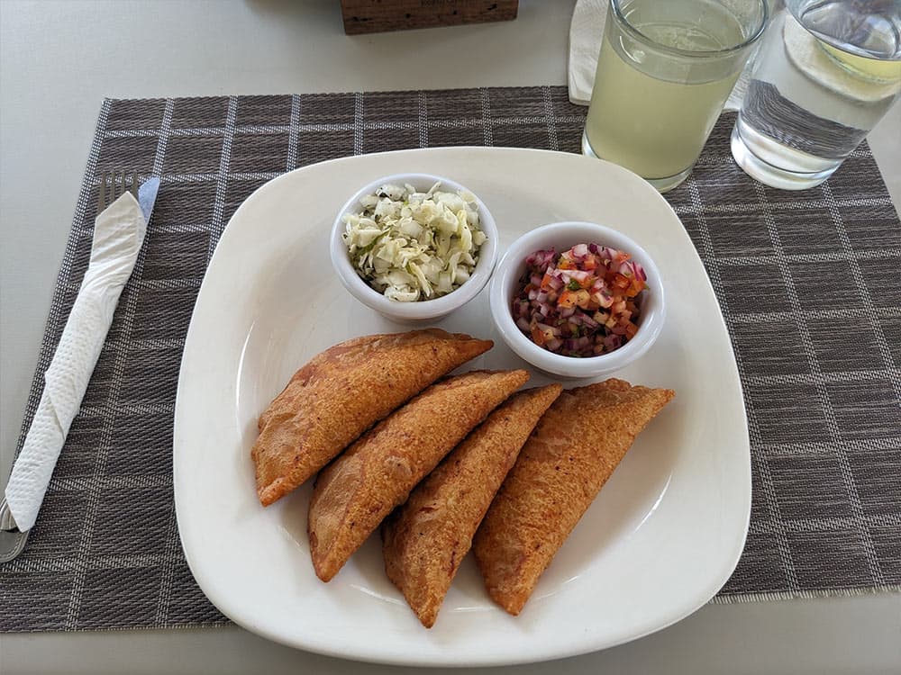 A plate of four golden fried empanadas served with two small bowls of coleslaw and salsa, accompanied by a glass of lemon drink.