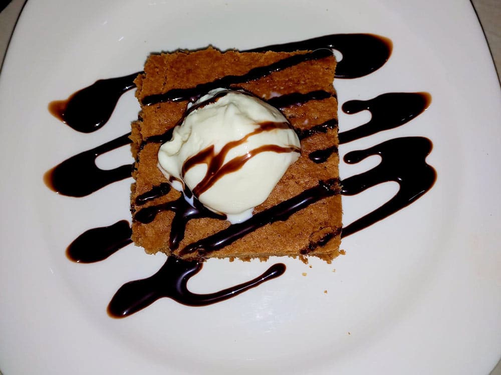 A square piece of dessert drizzled with chocolate sauce and topped with a scoop of vanilla ice cream.