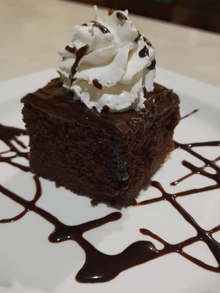 A slice of chocolate cake topped with whipped cream and drizzled with chocolate sauce on a white plate.