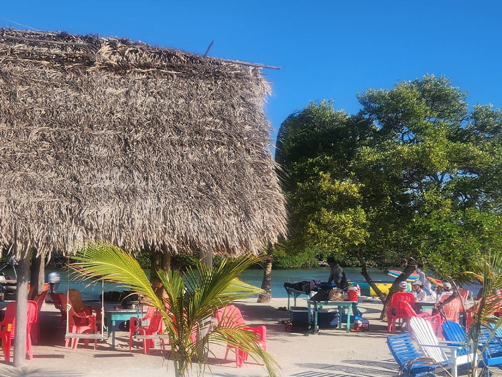A beachside scene featuring a thatched roof structure, colorful chairs, and people gathered near the water.