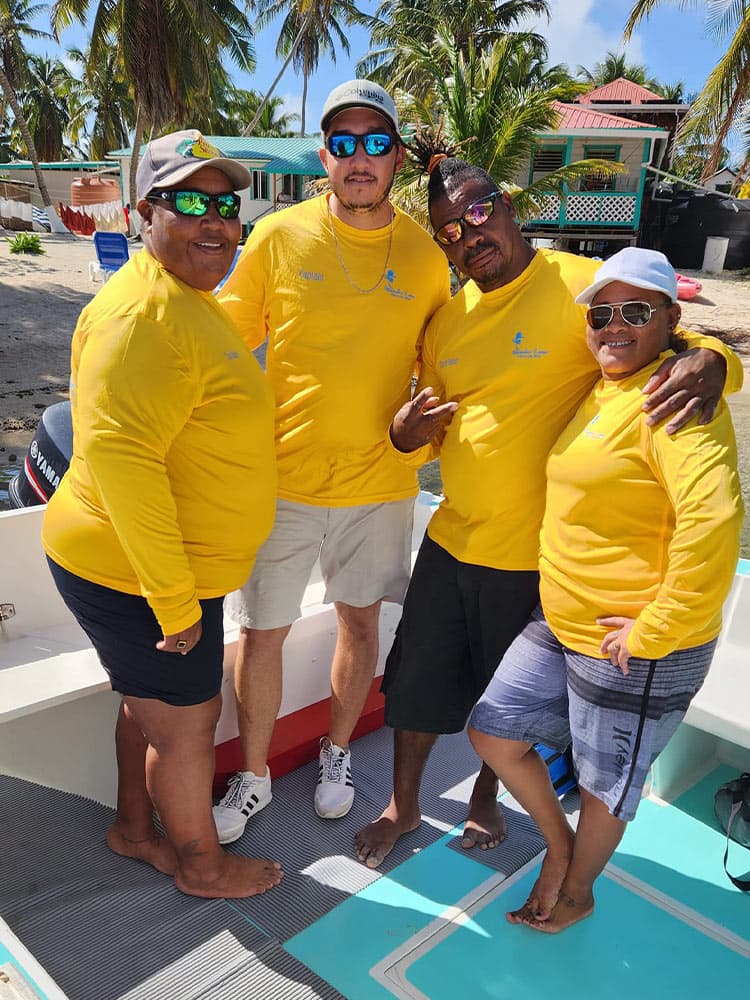 Four people wearing matching yellow shirts pose together on a boat with palm trees and colorful buildings in the background.