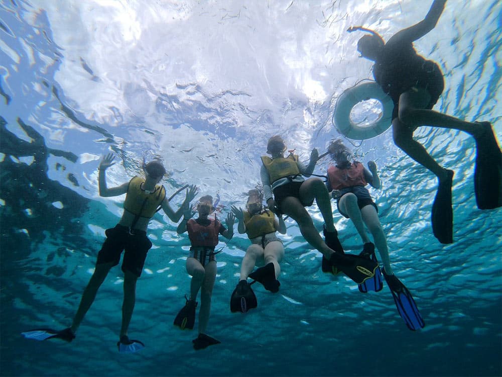 A group of six people wearing snorkeling gear and life jackets swim underwater while one person floats above them.