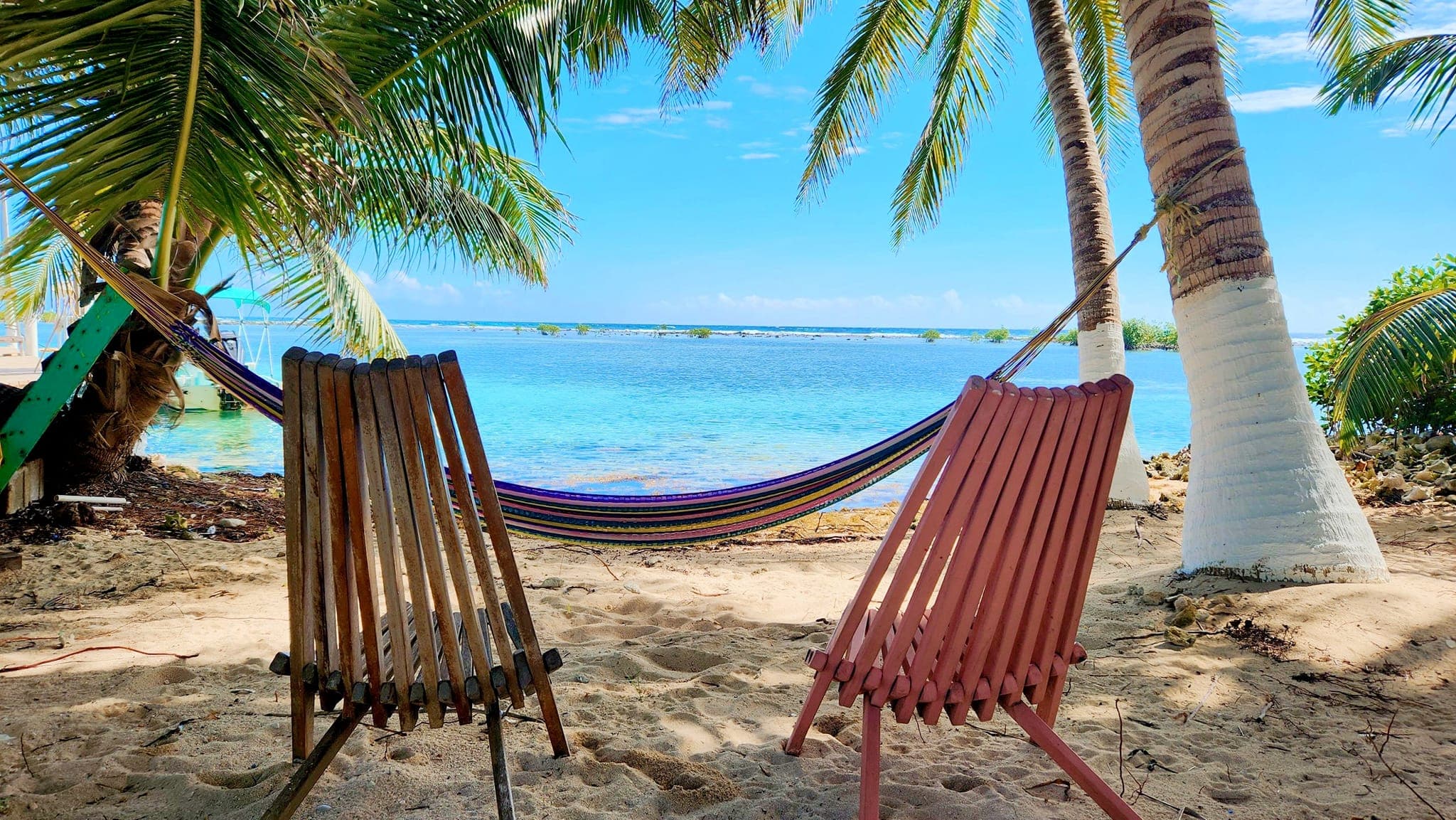 Two colorful chairs beside a hammock on a sandy beach with palm trees and calm blue water.