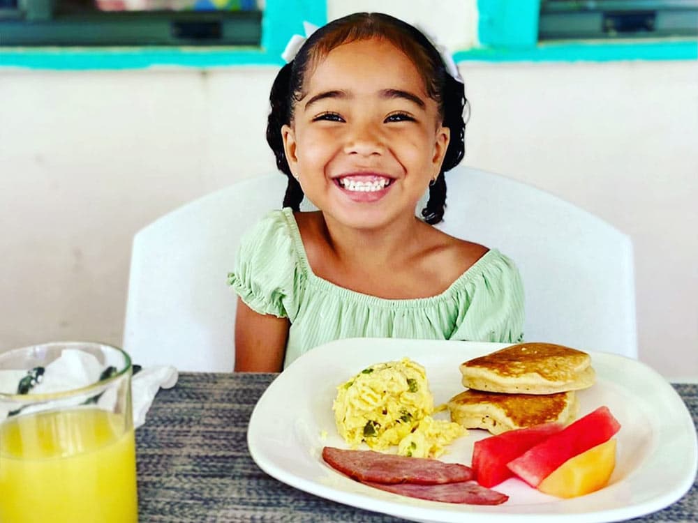 A smiling girl in a green shirt enjoys a plate of breakfast with pancakes, scrambled eggs, fruit, and a glass of juice.