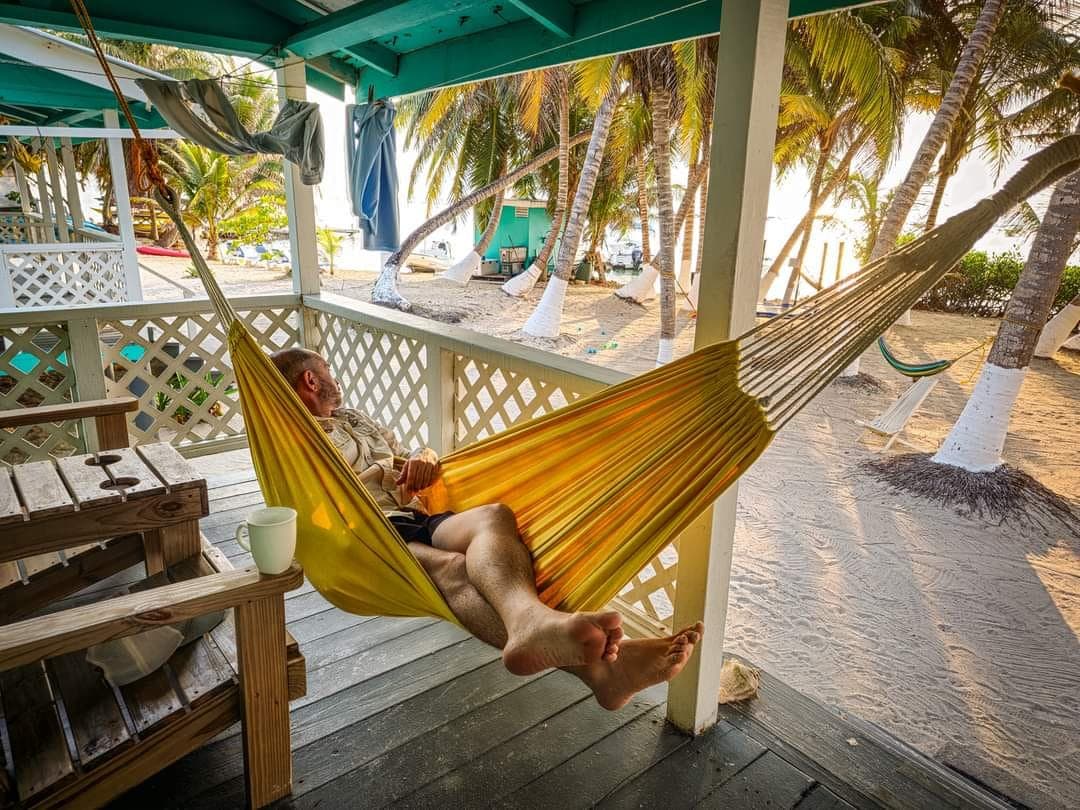 A person relaxes in a yellow hammock on a porch surrounded by palm trees.