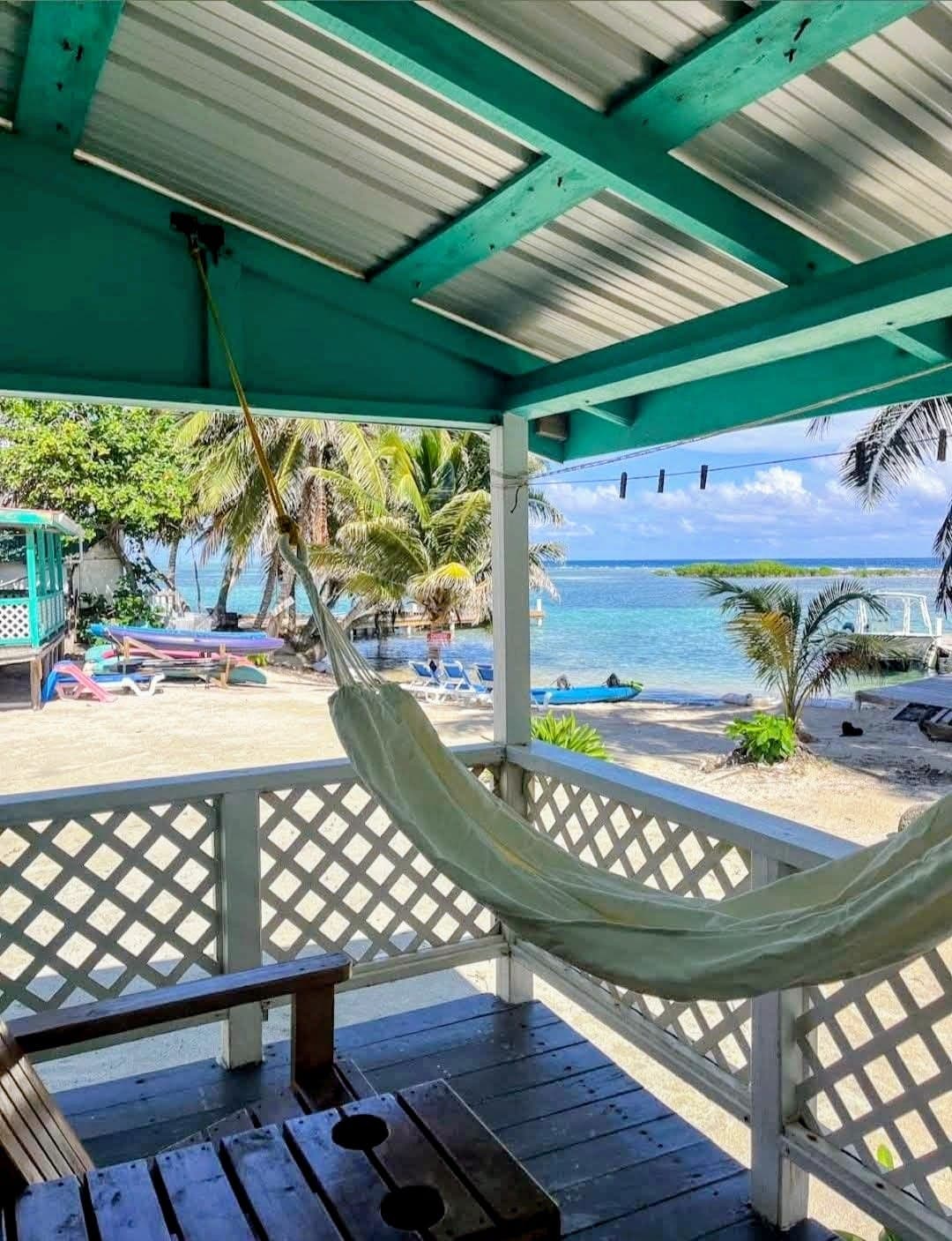 A hammock hangs in a shaded porch overlooking a sandy beach and turquoise waters.