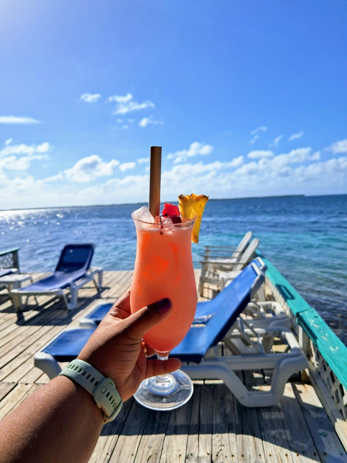A hand holds a colorful cocktail with a straw and fruit garnish, set against a backdrop of a sunny beach and ocean.