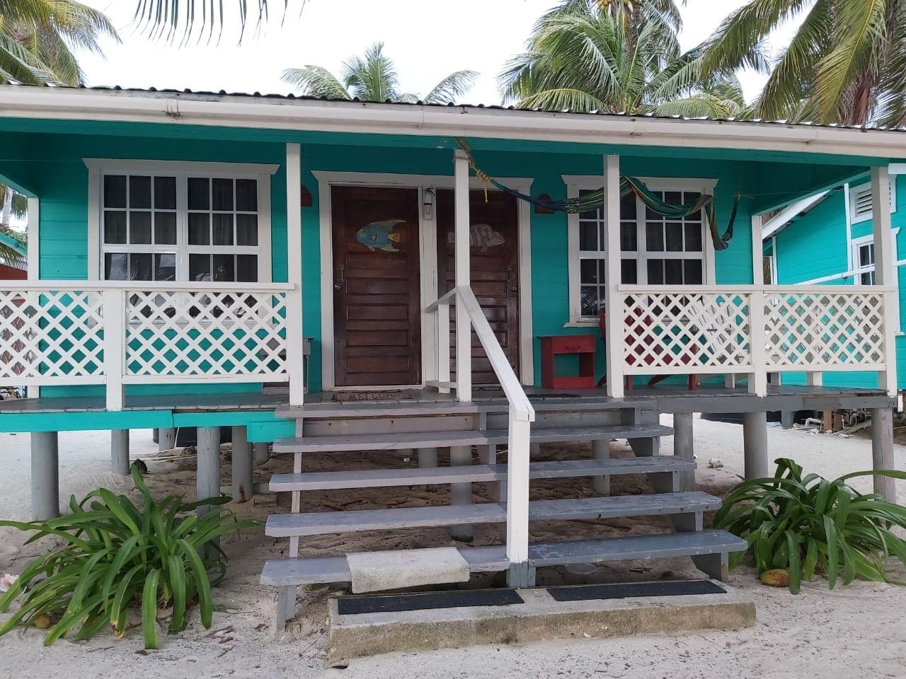 A turquoise beach house with stairs leading to a porch, surrounded by palm trees.