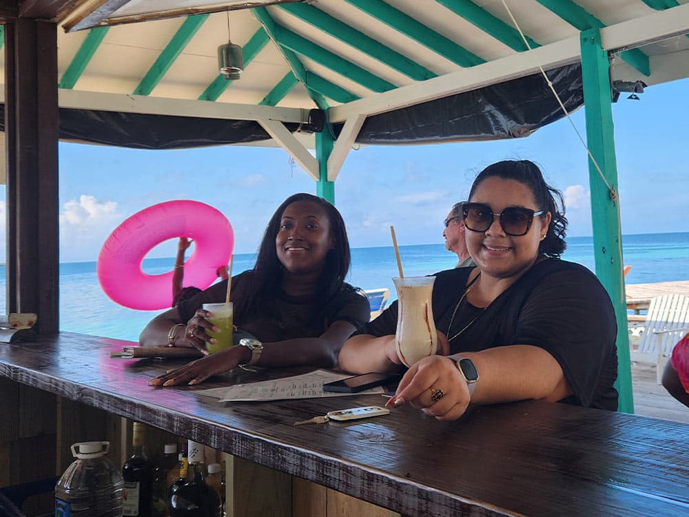 Two women smile while sitting at a beach bar, each holding a tropical drink, with a peaceful ocean backdrop.