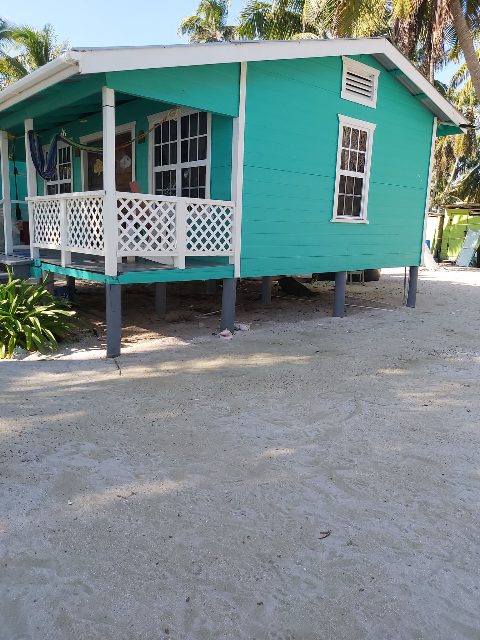 A teal beach house with white railings stands on stilts in a sandy area surrounded by palm trees.