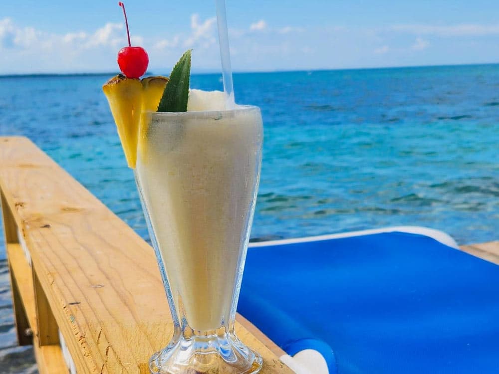 A tropical cocktail garnished with a cherry, pineapple, and a straw sits on a wooden table by the ocean.