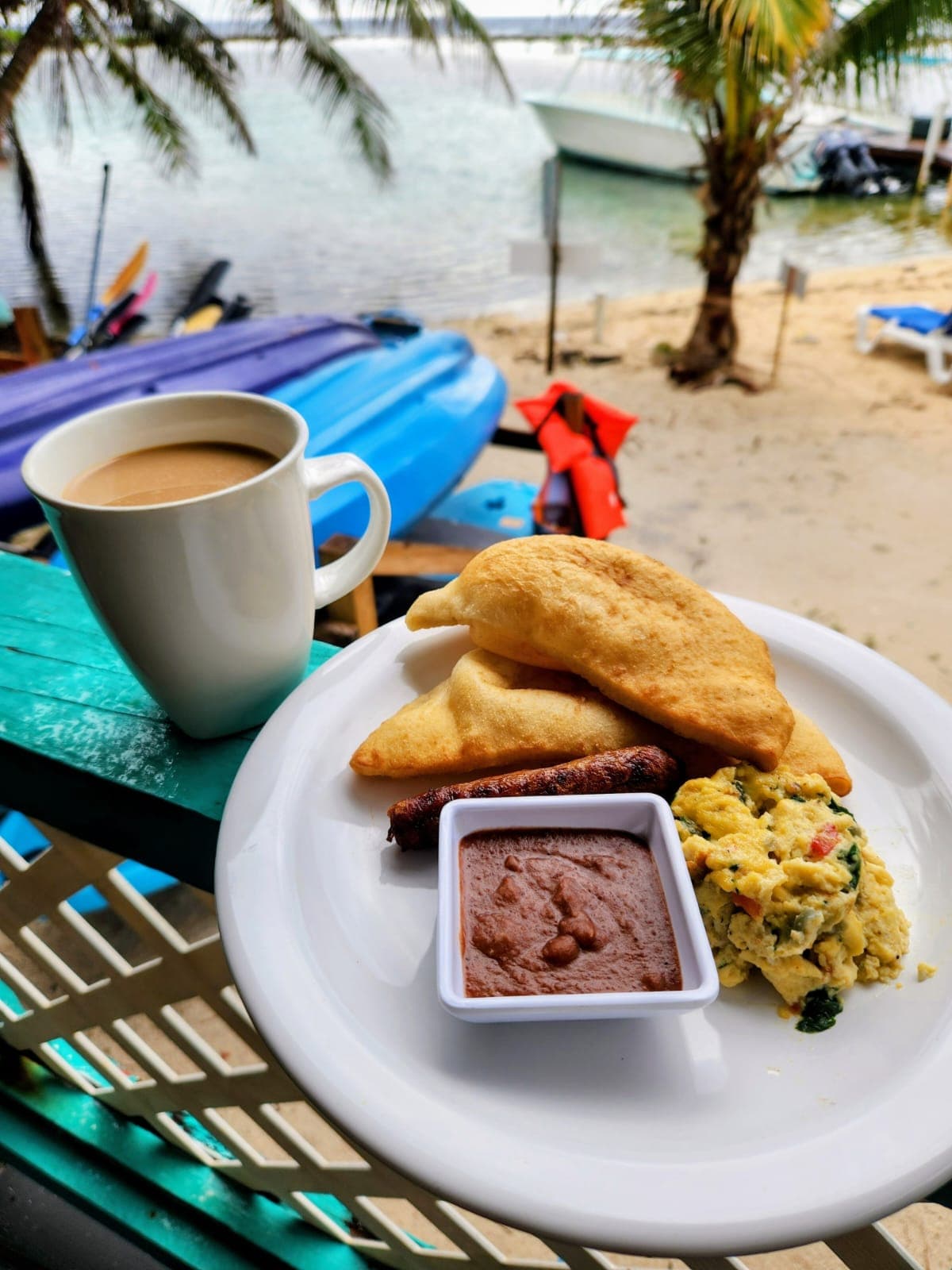A plate of scrambled eggs, fried dough, sausage, and sauce, accompanied by a cup of coffee, with a beach view in the background.