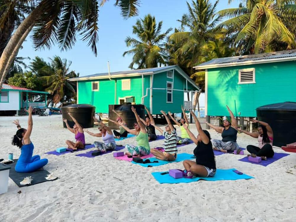 A group of women practicing yoga on a sandy beach in front of colorful cabins, surrounded by palm trees.