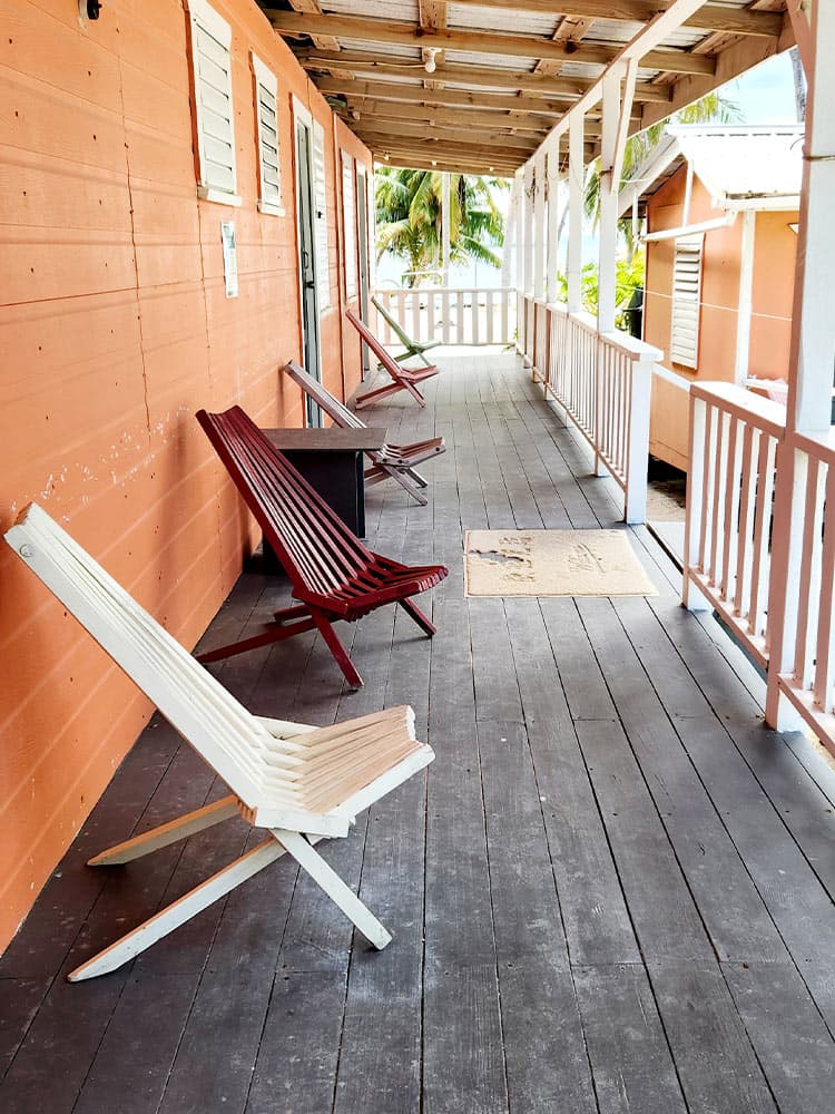 A wooden porch with two red and one white lounge chairs, and a small table, surrounded by palm trees.