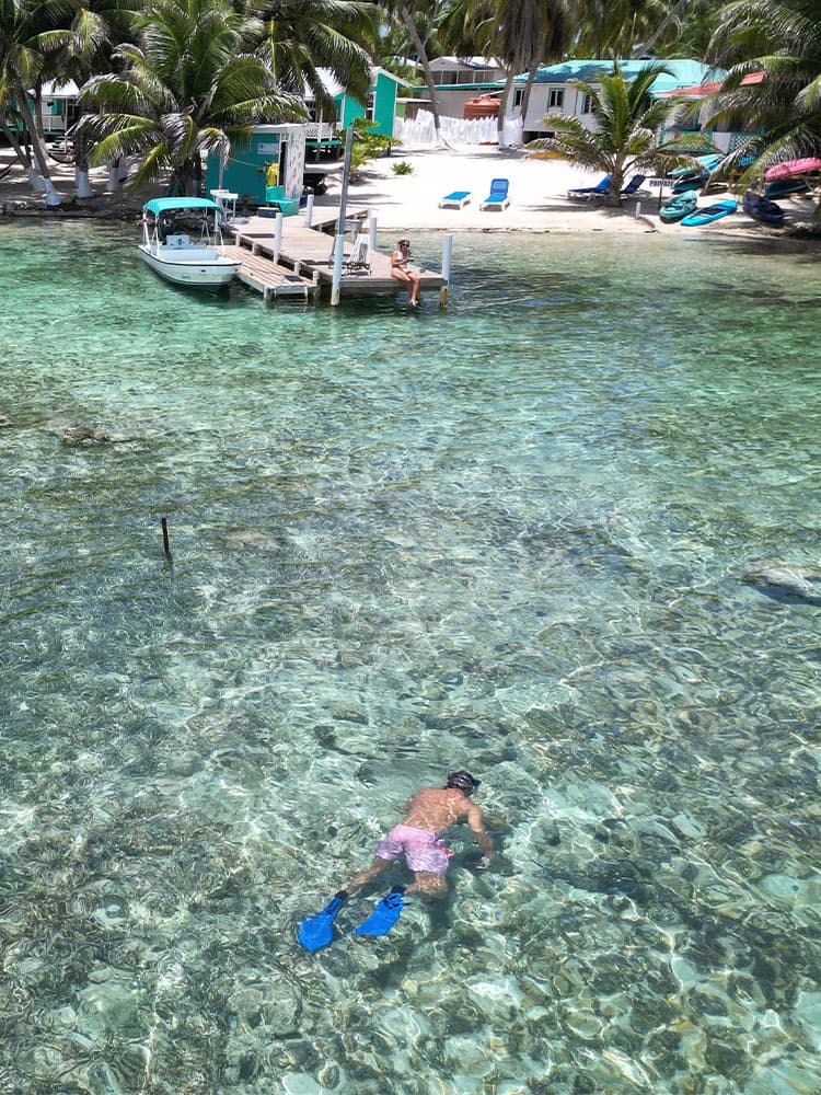 A swimmer in blue fins explores clear water near a dock with a boat and beach chairs in the background.