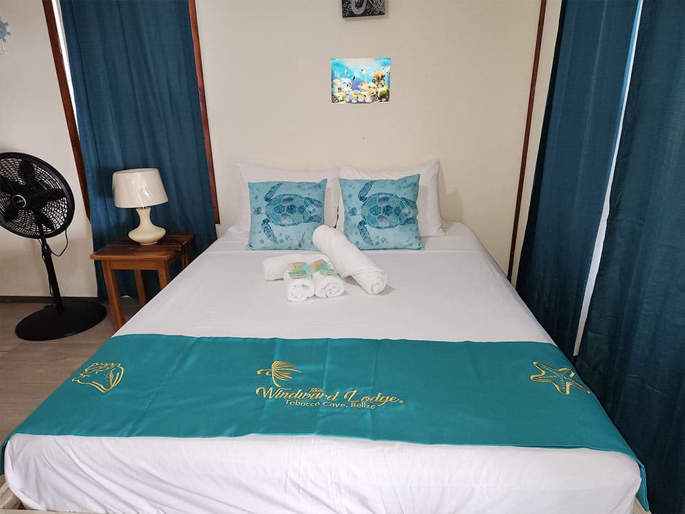 A neatly made bed with turquoise accents, decorative pillows, and towels, set against a backdrop of blue curtains and a fan.