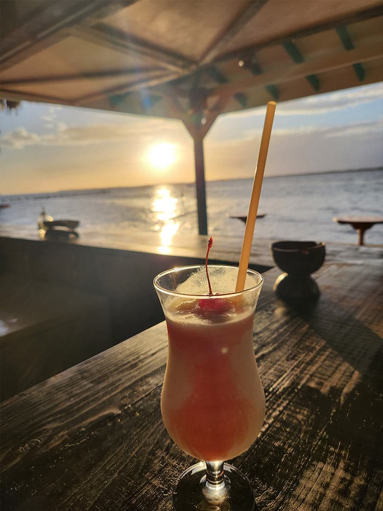 A tropical cocktail with a cherry sits on a table, framed by a sunset over the water.