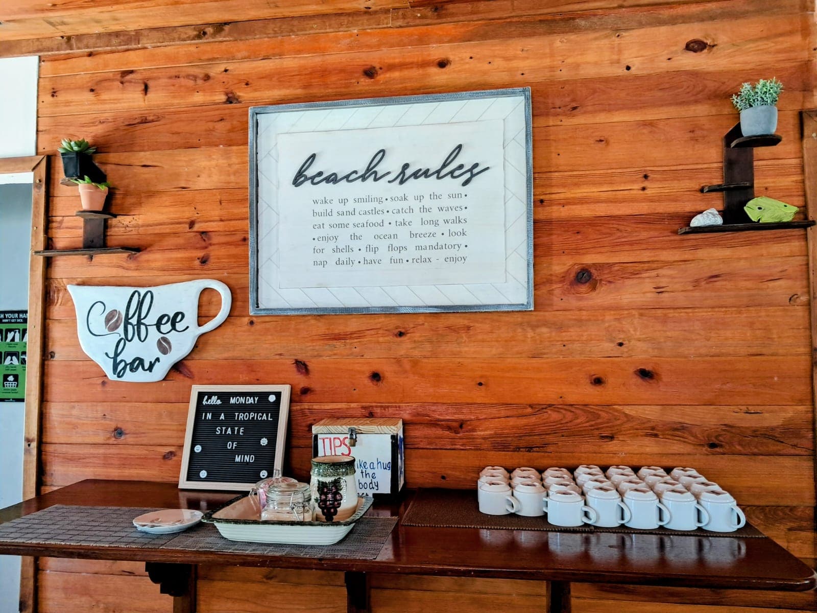 A rustic coffee bar with a sign displaying beach rules, surrounded by small plants and neatly arranged mugs.